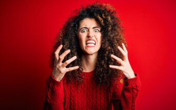 A person with curly hair and a nose ring is in front of a red background. They are wearing a red sweater and have a frustrated expression, with their hands raised and fingers spread apart.