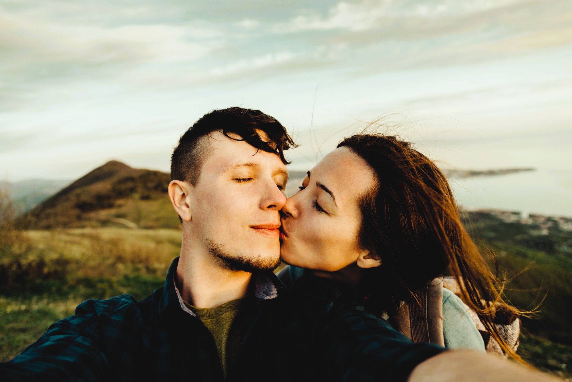 A couple takes a selfie on a hilltop, with the woman kissing the man's cheek. They are surrounded by scenic views of distant hills and a body of water under a cloudy sky. The moment captures a sense of affection and adventure.