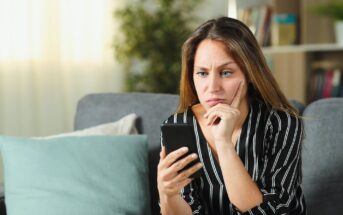 A woman with long brown hair sits on a gray couch in a living room, looking at her smartphone with a thoughtful expression. She rests her chin on her hand and wears a black and white striped shirt. There are cushions and a blurred bookshelf behind her.