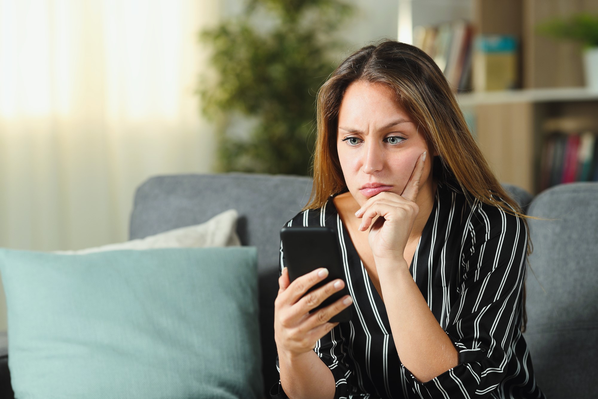 A woman with long brown hair sits on a gray couch in a living room, looking at her smartphone with a thoughtful expression. She rests her chin on her hand and wears a black and white striped shirt. There are cushions and a blurred bookshelf behind her.