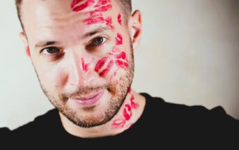 Man with a short beard smiling at the camera, wearing a black shirt. His face and neck are covered with multiple red lipstick marks, suggesting kisses. The background is a light, neutral color.