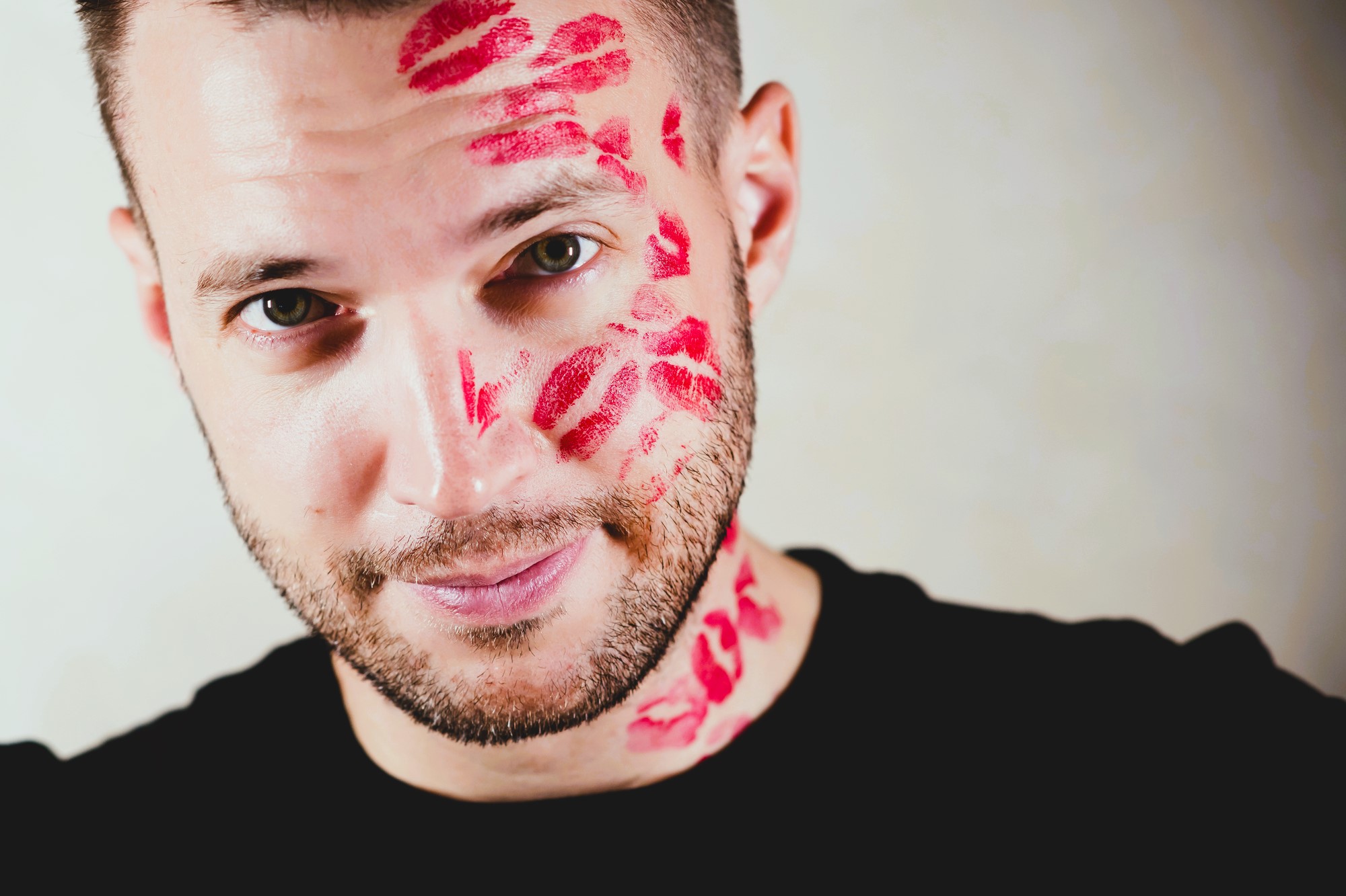 Man with a short beard smiling at the camera, wearing a black shirt. His face and neck are covered with multiple red lipstick marks, suggesting kisses. The background is a light, neutral color.