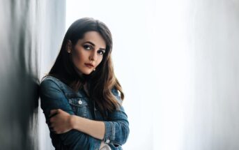 Woman with long brown hair and a serious expression, wearing a denim jacket. She is leaning against a light-colored wall with her arms crossed, looking towards the camera. The background is bright and softly blurred.