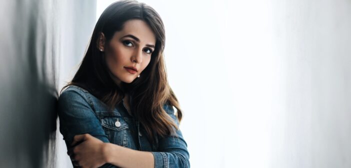 Woman with long brown hair and a serious expression, wearing a denim jacket. She is leaning against a light-colored wall with her arms crossed, looking towards the camera. The background is bright and softly blurred.