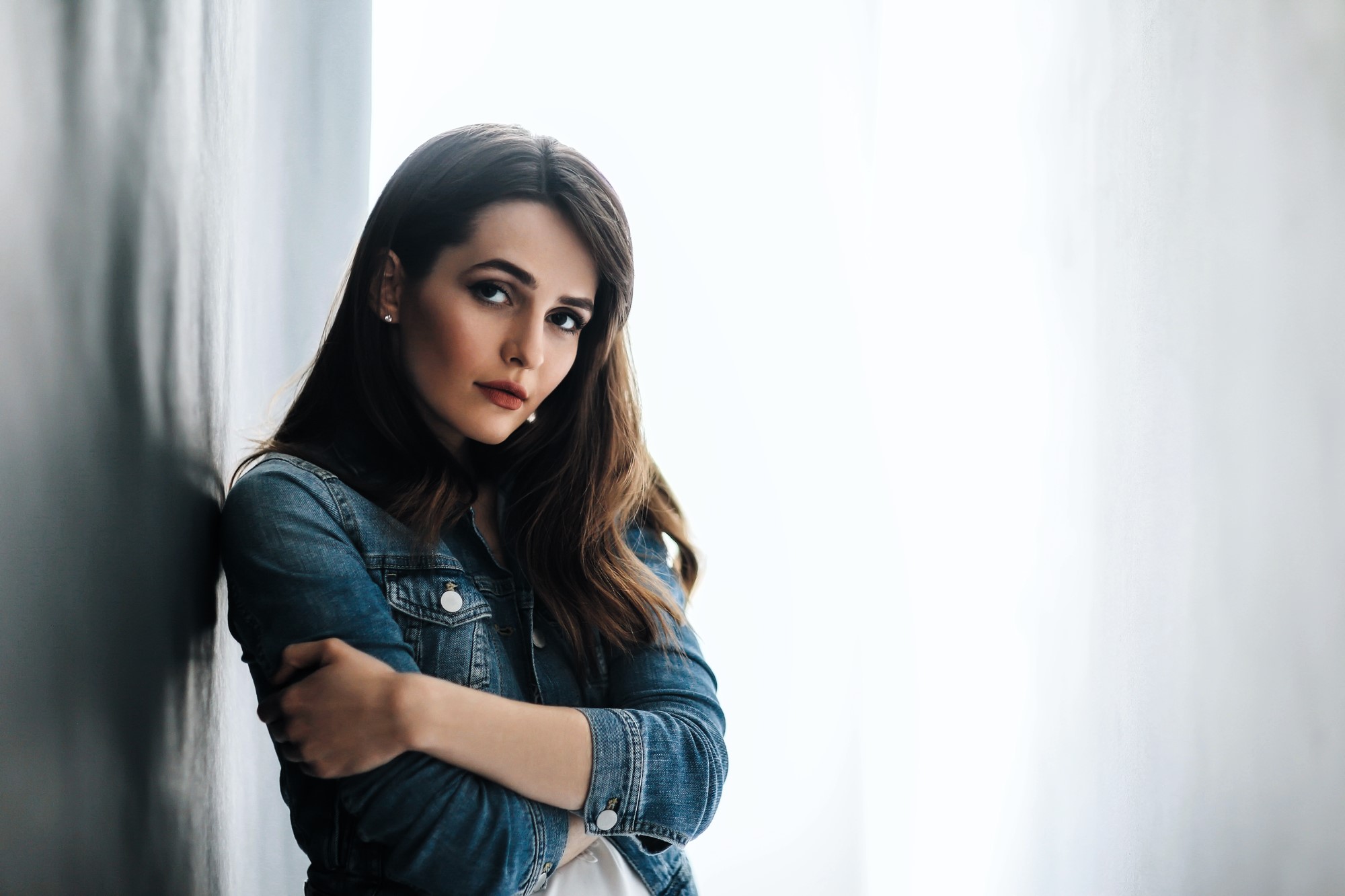 Woman with long brown hair and a serious expression, wearing a denim jacket. She is leaning against a light-colored wall with her arms crossed, looking towards the camera. The background is bright and softly blurred.