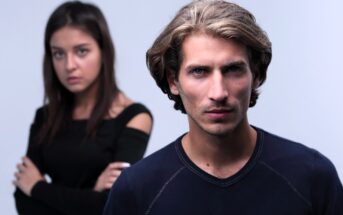 A man with shoulder-length brown hair and a serious expression stands in the foreground, wearing a dark shirt. In the blurred background, a woman with long brown hair, wearing a black top, looks on with her arms crossed.