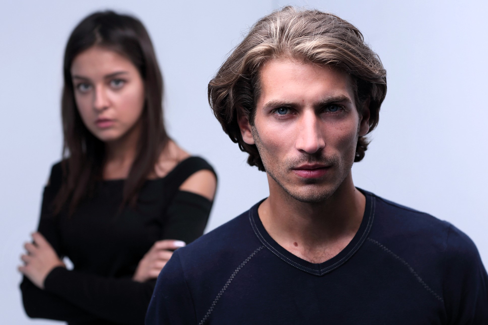 A man with shoulder-length brown hair and a serious expression stands in the foreground, wearing a dark shirt. In the blurred background, a woman with long brown hair, wearing a black top, looks on with her arms crossed.