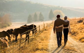 A couple walks arm-in-arm on a rural path beside a fenced field with horses grazing. The scene is set in the warm glow of a setting sun, with hills and trees in the background, creating a peaceful countryside atmosphere.