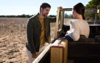 A man and a woman are interacting by a parked open-top vehicle in a dry, open field. The man is wearing a green jacket and leaning on the car door. The woman, in a white sweater, is seated inside with a suitcase next to her. Trees are in the background.