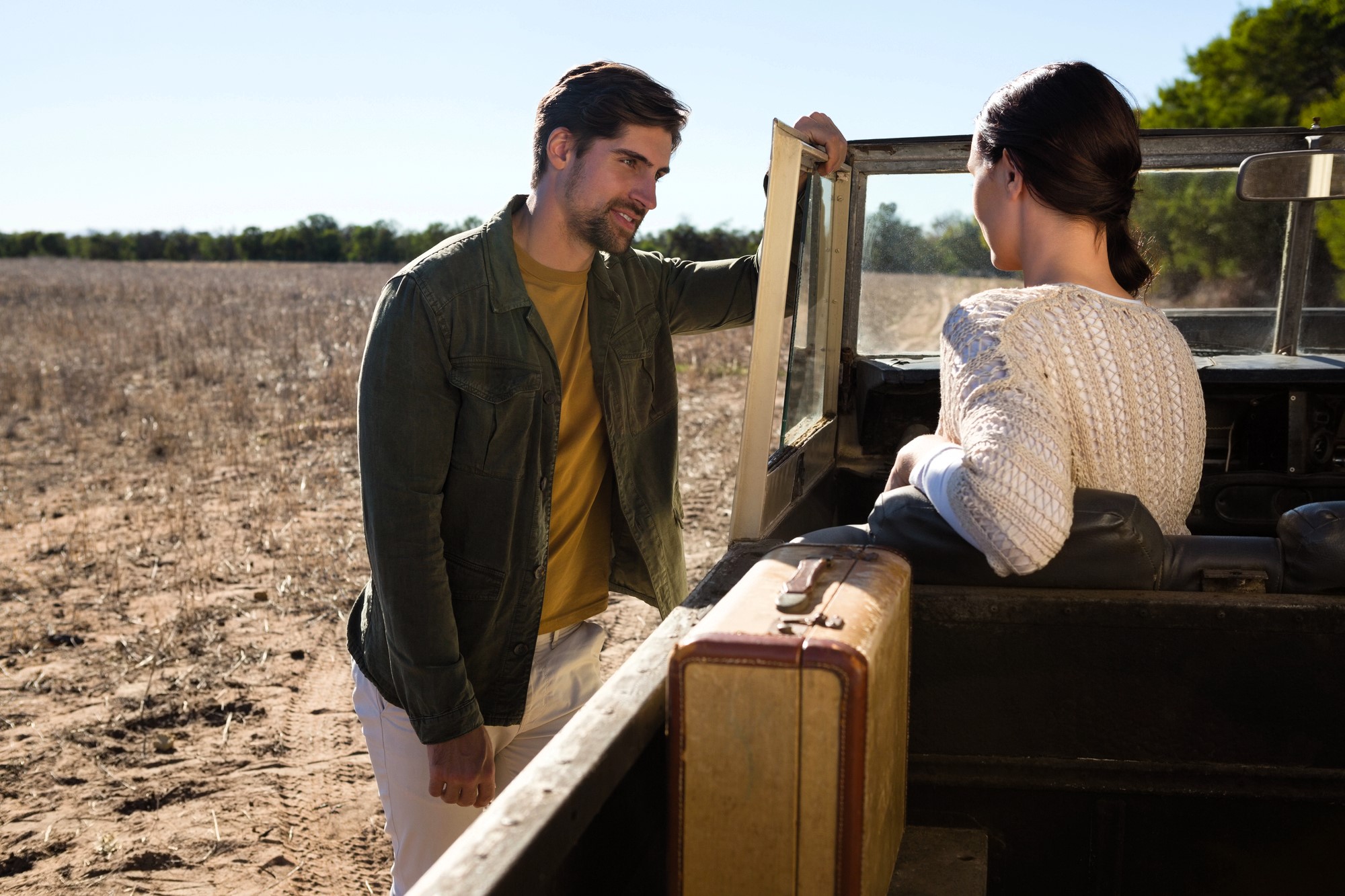 A man and a woman are interacting by a parked open-top vehicle in a dry, open field. The man is wearing a green jacket and leaning on the car door. The woman, in a white sweater, is seated inside with a suitcase next to her. Trees are in the background.