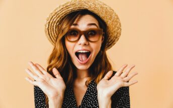 A woman wearing a straw hat and large sunglasses is posing excitedly with wide open mouth and hands raised near her face. She has long brown hair and is wearing a black polka dot dress. The background is a solid peach color.