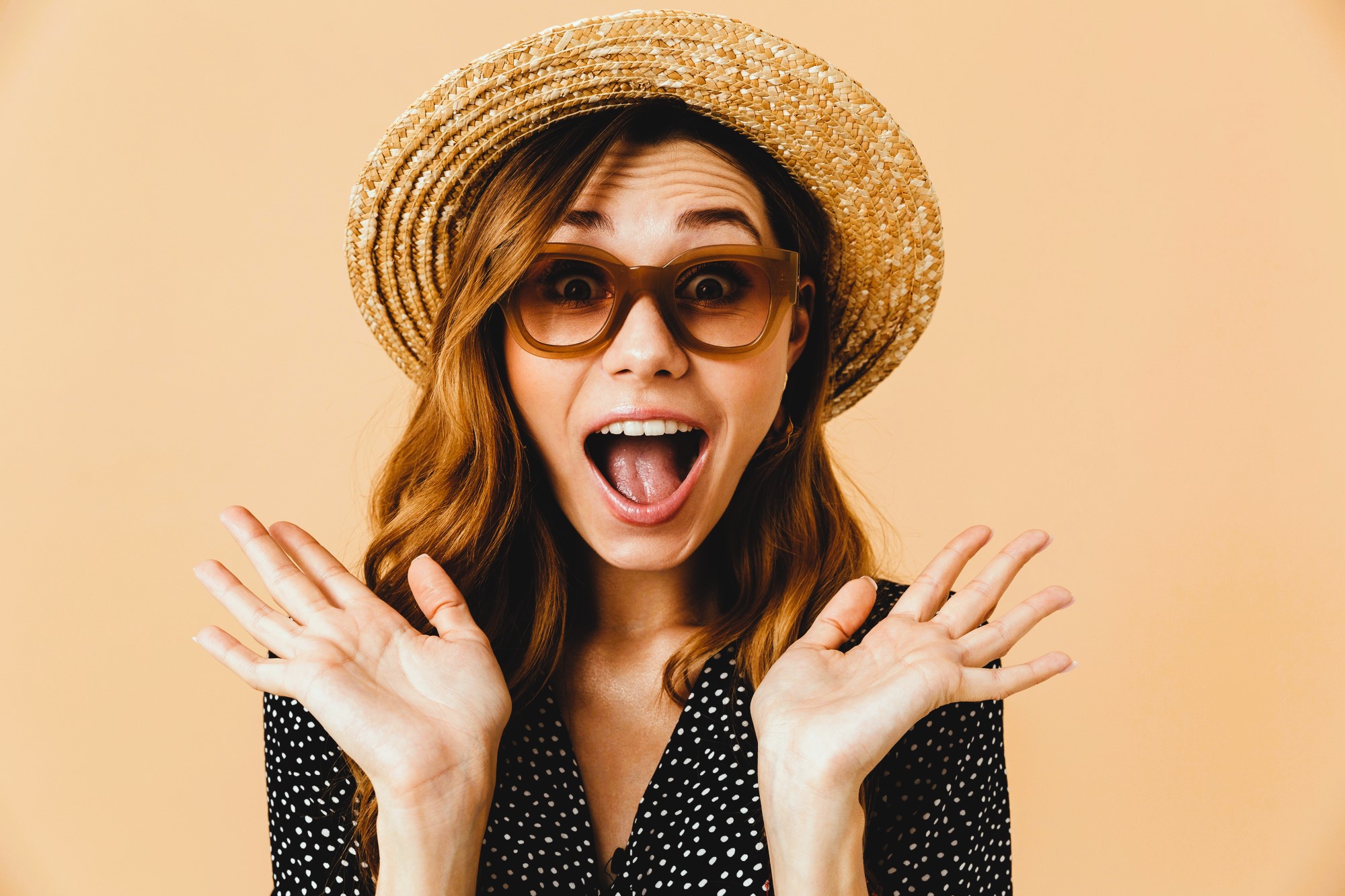 A woman wearing a straw hat and large sunglasses is posing excitedly with wide open mouth and hands raised near her face. She has long brown hair and is wearing a black polka dot dress. The background is a solid peach color.