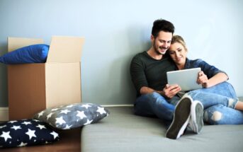 A couple is sitting on the floor leaning against a wall, looking at a tablet. They appear relaxed and happy. There are star-patterned cushions and a cardboard box nearby, suggesting they've just moved in or are unpacking.