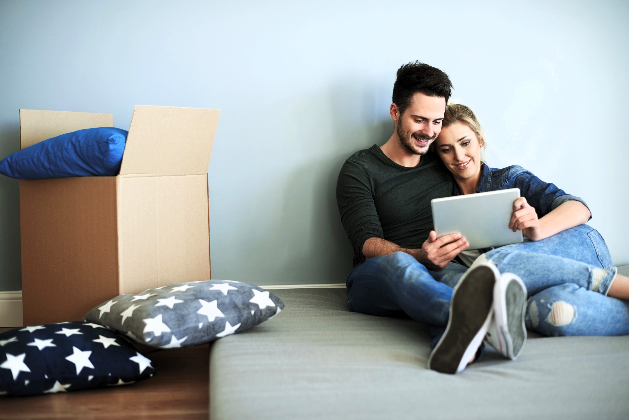 A couple is sitting on the floor leaning against a wall, looking at a tablet. They appear relaxed and happy. There are star-patterned cushions and a cardboard box nearby, suggesting they've just moved in or are unpacking.