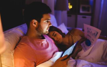 A man in a pink shirt is sitting in bed using a tablet, with charts and graphs on papers beside him. A woman is sleeping next to him. The room is dimly lit, suggesting it is nighttime.