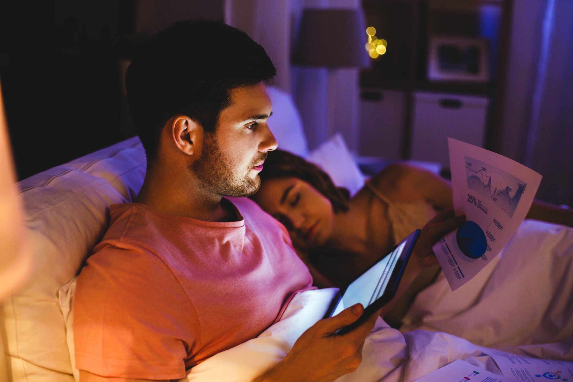 A man in a pink shirt is sitting in bed using a tablet, with charts and graphs on papers beside him. A woman is sleeping next to him. The room is dimly lit, suggesting it is nighttime.