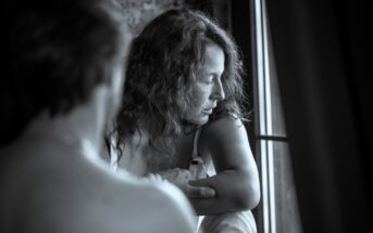 A black and white image shows a woman with curly hair sitting by a window, looking contemplatively outside. A blurred figure, seen from behind, is in the foreground, partially obscuring the view.