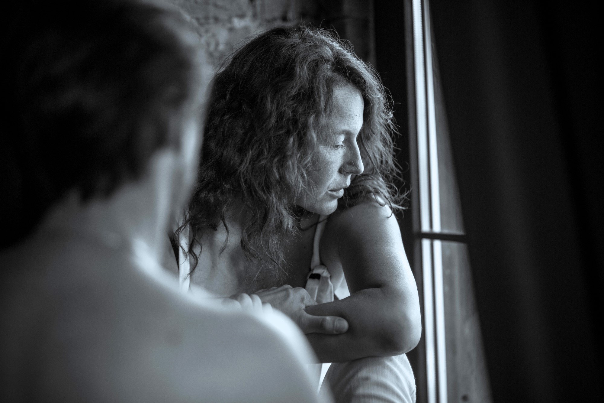 A black and white image shows a woman with curly hair sitting by a window, looking contemplatively outside. A blurred figure, seen from behind, is in the foreground, partially obscuring the view.