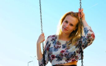 A woman with blonde hair smiles while sitting on a swing against a clear blue sky. She is wearing a long-sleeve floral top and holding the swing chains with both hands. A streetlamp is visible in the background.