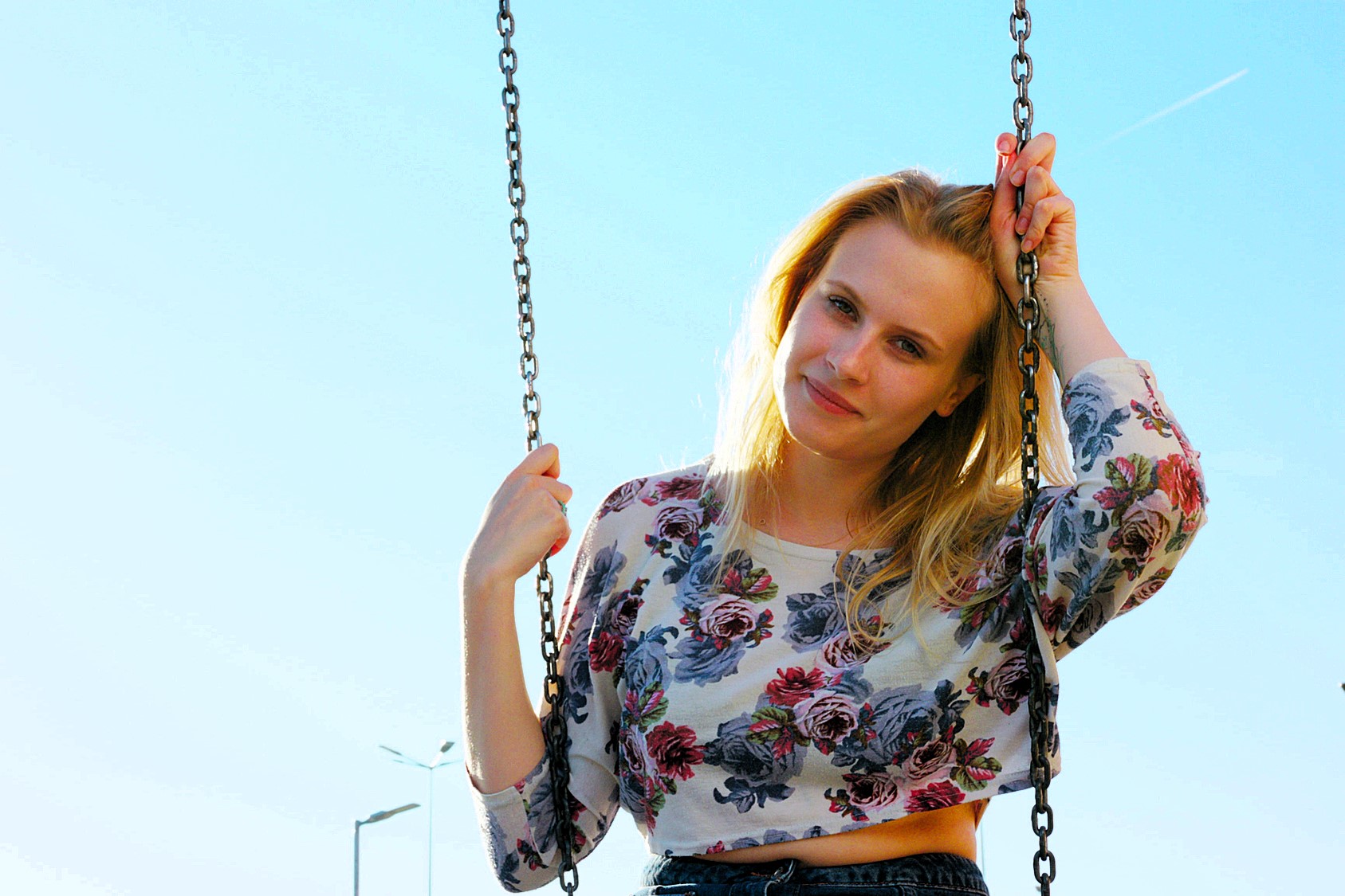 A woman with blonde hair smiles while sitting on a swing against a clear blue sky. She is wearing a long-sleeve floral top and holding the swing chains with both hands. A streetlamp is visible in the background.