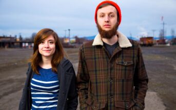 A woman and a man stand outdoors on a cloudy day. The woman on the left has shoulder-length brown hair and a striped shirt, while the man on the right has a beard and wears an orange beanie and brown coat. A barren landscape is in the background.