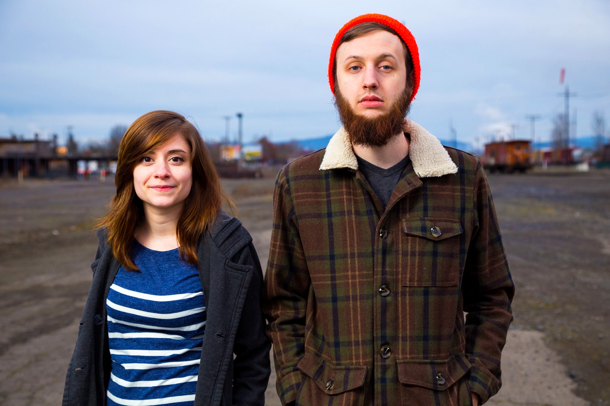 A woman and a man stand outdoors on a cloudy day. The woman on the left has shoulder-length brown hair and a striped shirt, while the man on the right has a beard and wears an orange beanie and brown coat. A barren landscape is in the background.