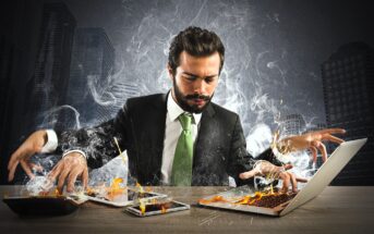 A man in a suit sits at a desk in a city office. He uses multiple devices, all emitting smoke and flames. The intense scene suggests stress and overload. The background features tall skyscrapers under a dark, stormy sky.