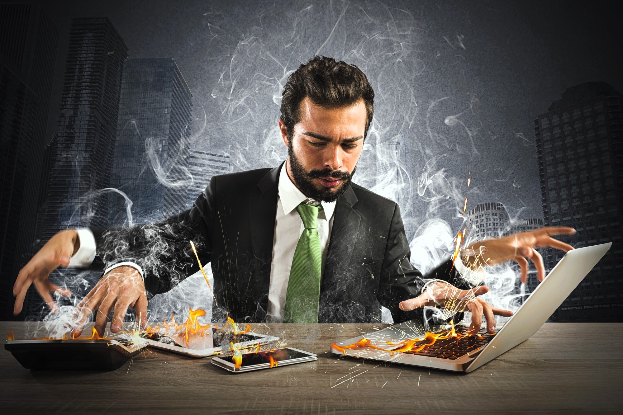 A man in a suit sits at a desk in a city office. He uses multiple devices, all emitting smoke and flames. The intense scene suggests stress and overload. The background features tall skyscrapers under a dark, stormy sky.