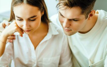 A young couple closely looks at something off-camera. The woman, with brown hair and a white blouse, rests her chin on her hand. The man, wearing a light shirt, leans in and gazes in the same direction, with his arm around her shoulders.