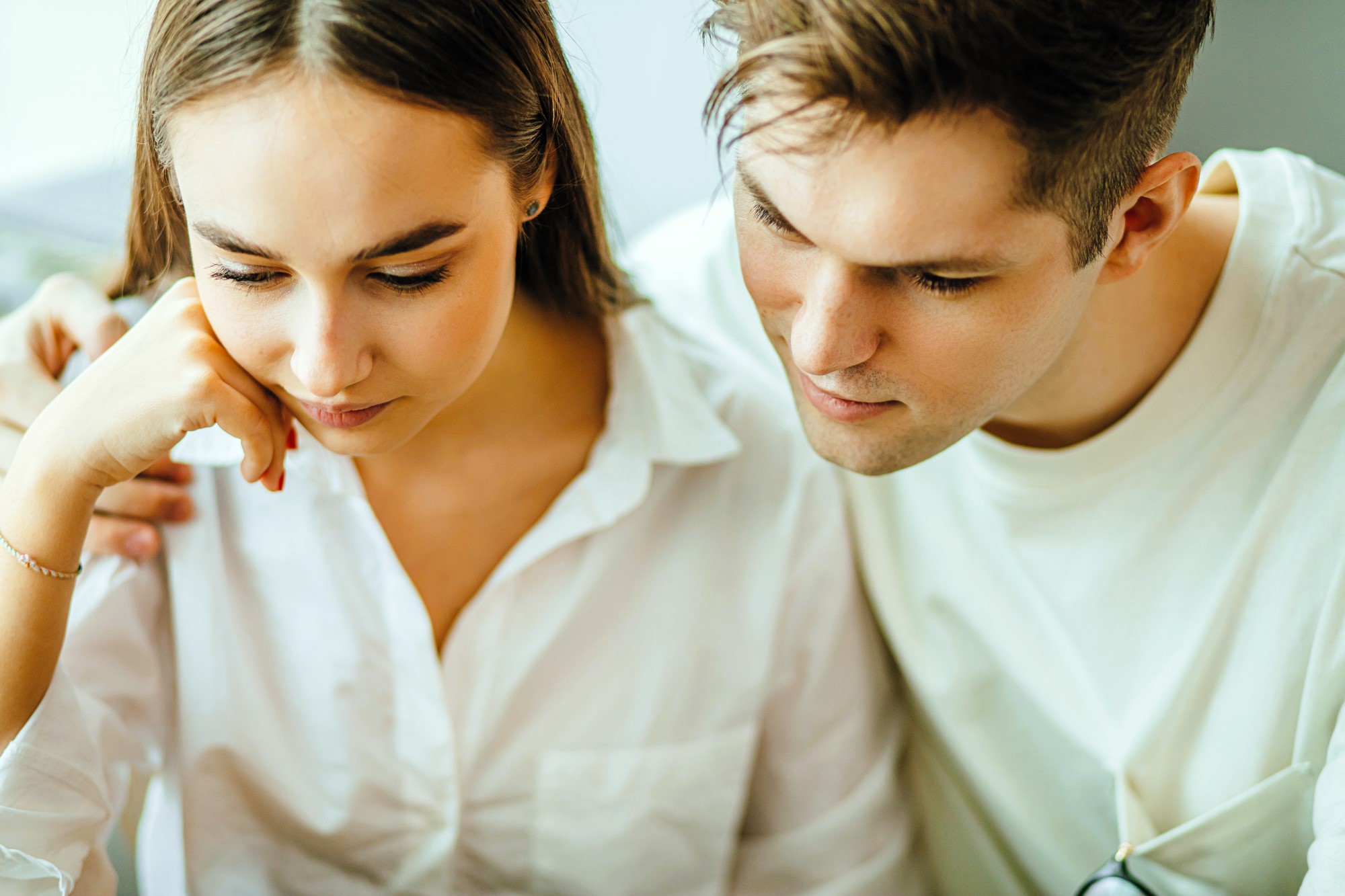 A young couple closely looks at something off-camera. The woman, with brown hair and a white blouse, rests her chin on her hand. The man, wearing a light shirt, leans in and gazes in the same direction, with his arm around her shoulders.