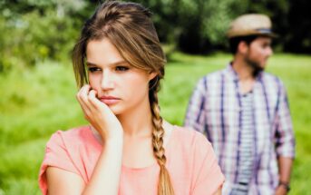 A woman with long braided hair and a pink top looks thoughtful, resting her chin on her hand. In the background, a man wearing a plaid shirt and a straw hat gazes into the distance. They are outdoors in a grassy area.