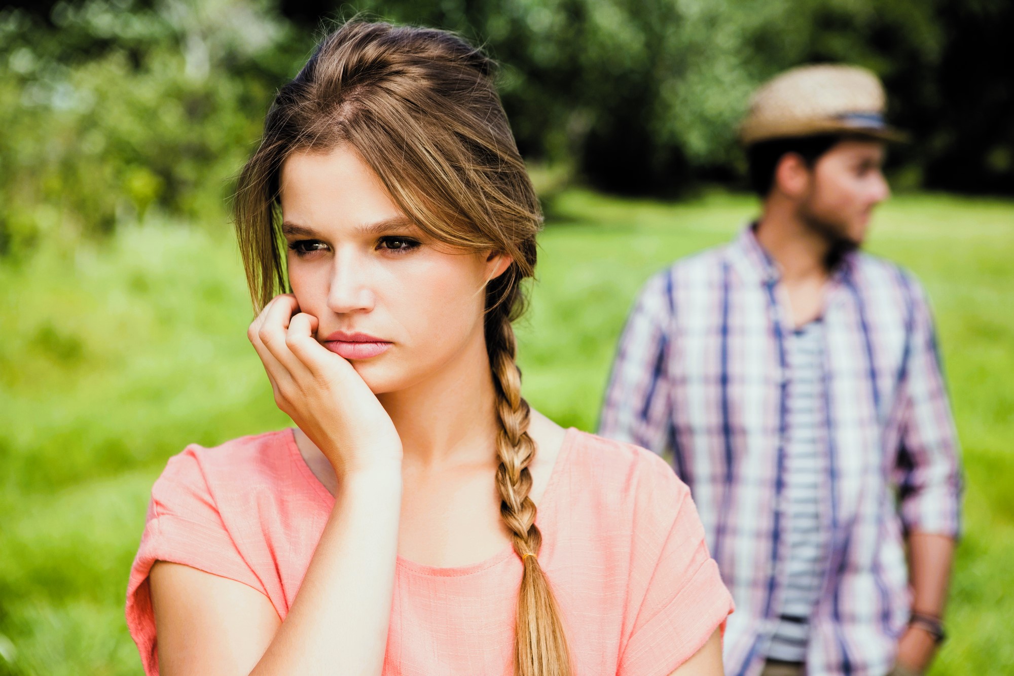 A woman with long braided hair and a pink top looks thoughtful, resting her chin on her hand. In the background, a man wearing a plaid shirt and a straw hat gazes into the distance. They are outdoors in a grassy area.