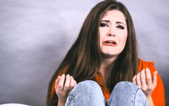 A woman with long brown hair sits against a gray background. She wears a red shirt and jeans, looking upset with tears on her face. Her hands are raised in a gesture of distress or frustration.