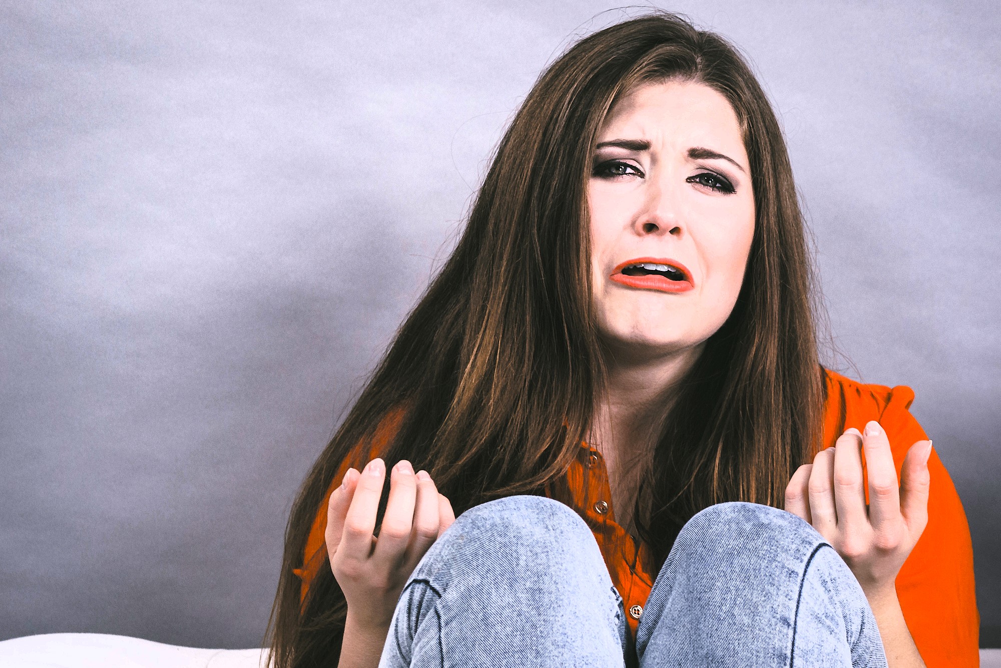 A woman with long brown hair sits against a gray background. She wears a red shirt and jeans, looking upset with tears on her face. Her hands are raised in a gesture of distress or frustration.