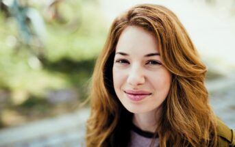 A young person with long, wavy hair smiles softly at the camera. They are outdoors with a blurred background of greenery and bicycles. The lighting is natural and soft.