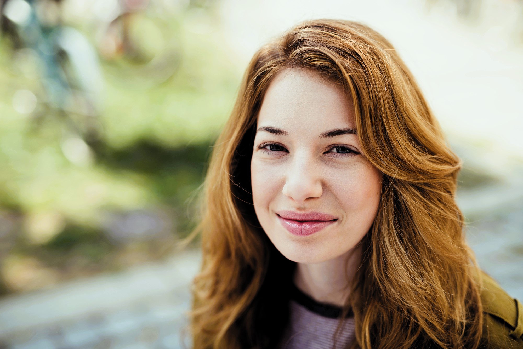 A young person with long, wavy hair smiles softly at the camera. They are outdoors with a blurred background of greenery and bicycles. The lighting is natural and soft.