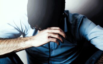 A man in a blue shirt sits with his head bowed, covering his face with one hand. The lighting casts a shadow, creating a somber atmosphere. His expression and posture convey a sense of distress or contemplation.