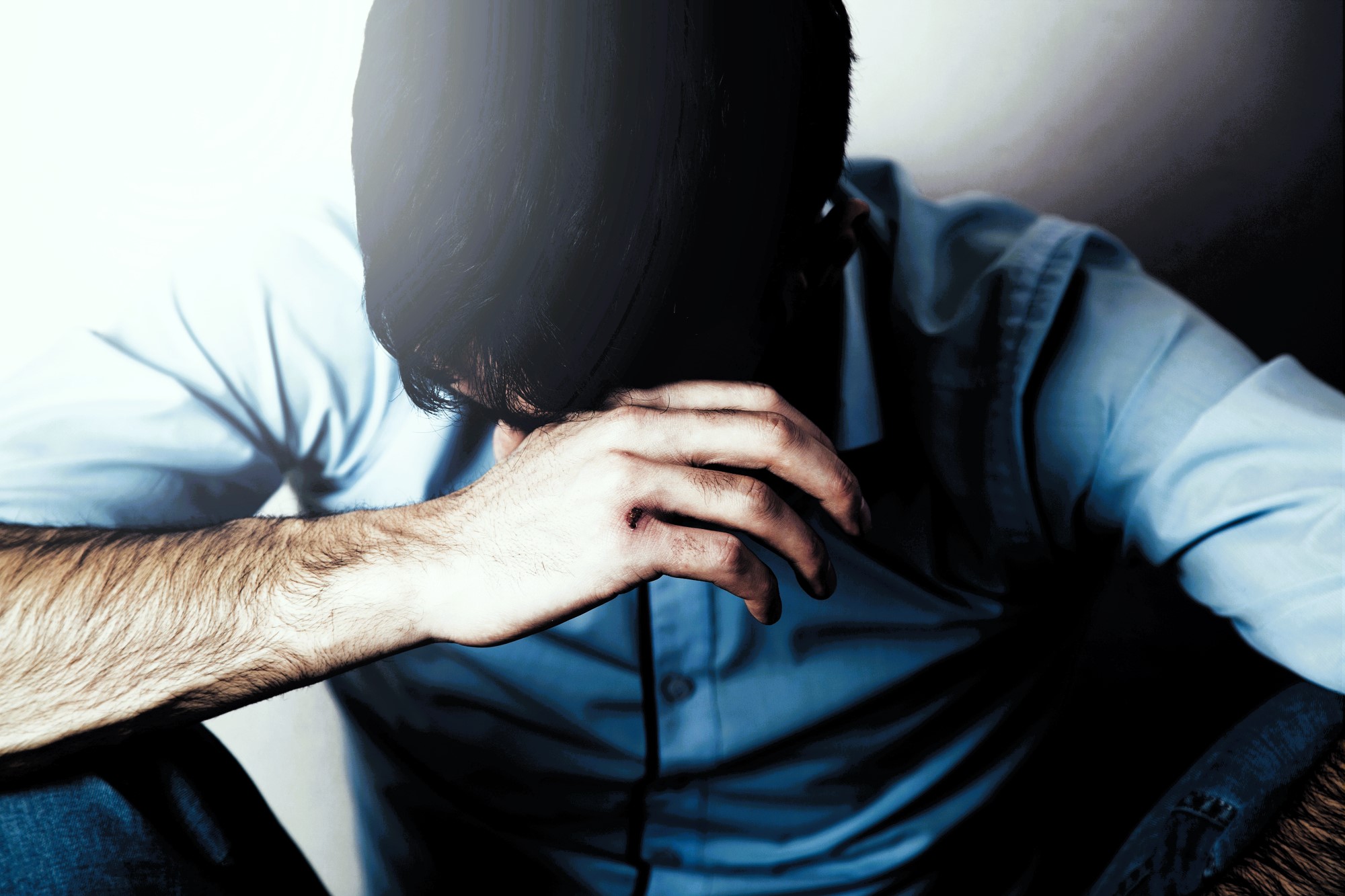 A man in a blue shirt sits with his head bowed, covering his face with one hand. The lighting casts a shadow, creating a somber atmosphere. His expression and posture convey a sense of distress or contemplation.