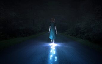 A person in a green dress walks alone on a dark road at night. Their feet and the path behind them are illuminated by a mysterious blue light, contrasting with the surrounding darkness.