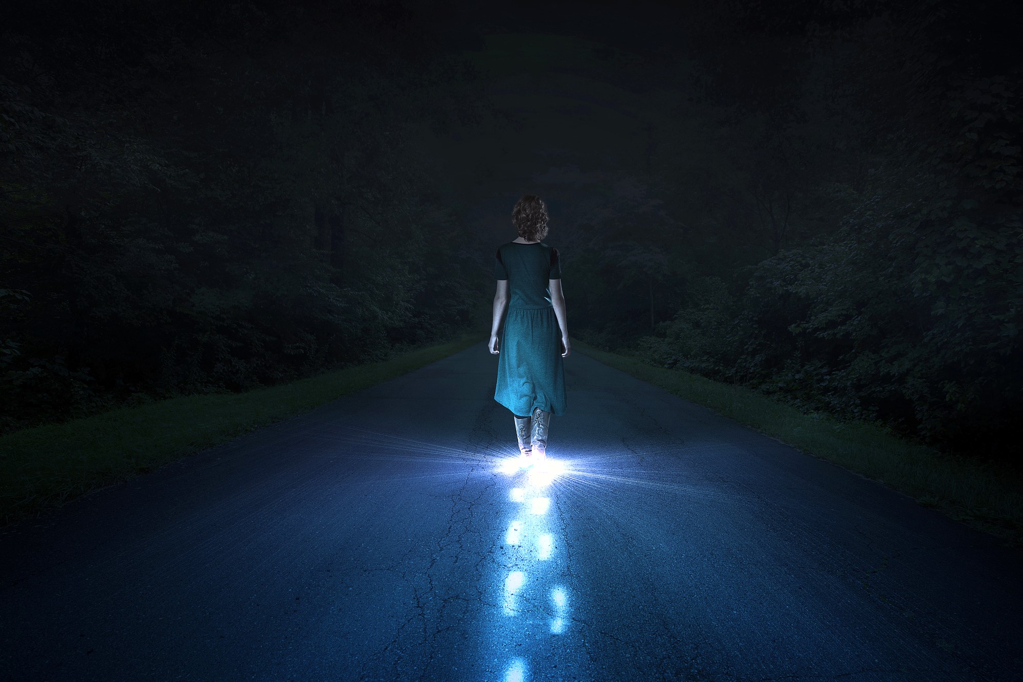 A person in a green dress walks alone on a dark road at night. Their feet and the path behind them are illuminated by a mysterious blue light, contrasting with the surrounding darkness.