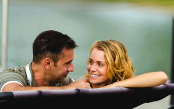 A man and a woman are relaxing in a hot tub outdoors. They are smiling at each other, appearing happy and enjoying their time together. The background is blurred, with greenery visible.