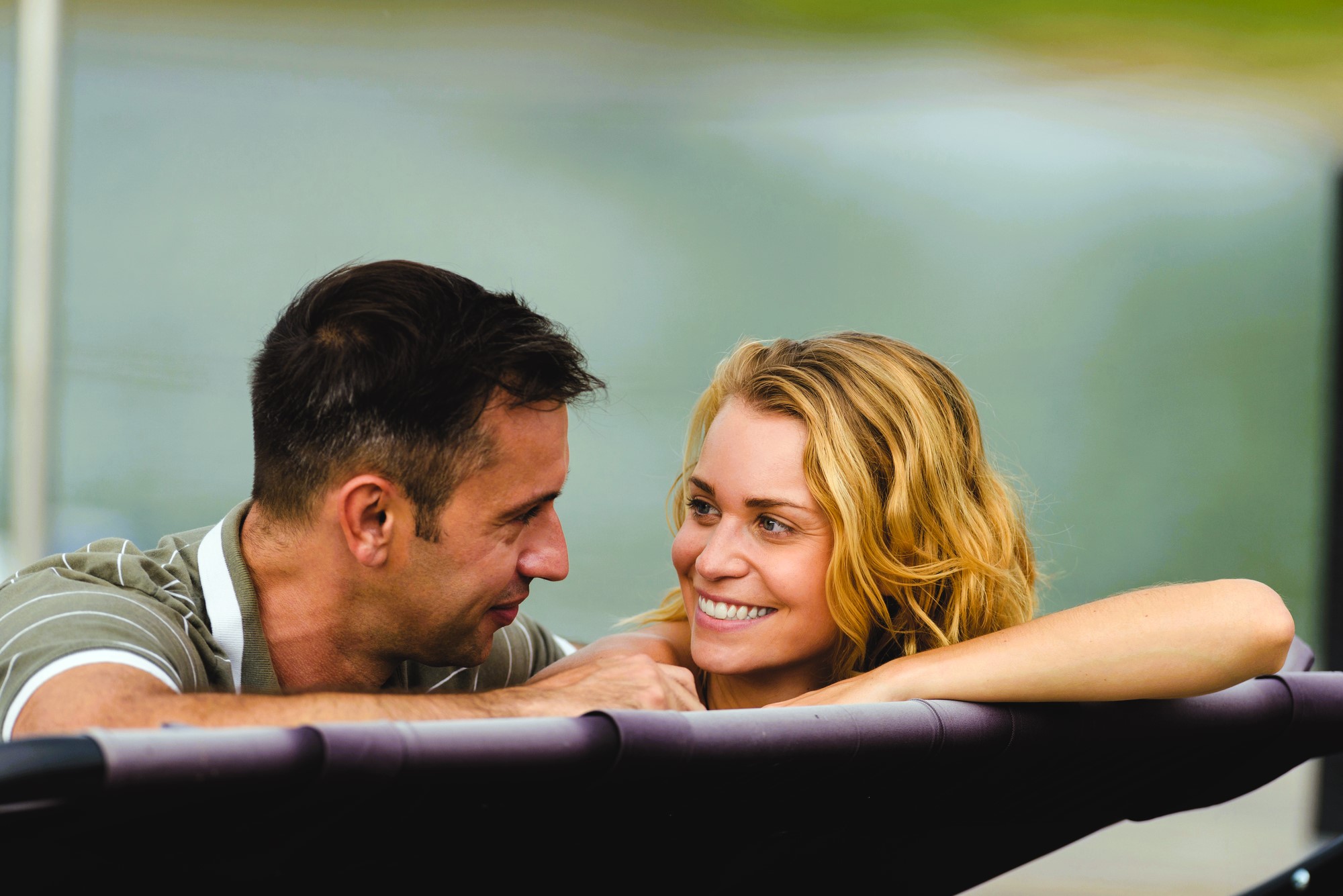 A man and a woman are relaxing in a hot tub outdoors. They are smiling at each other, appearing happy and enjoying their time together. The background is blurred, with greenery visible.