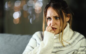 A woman wearing a striped turtleneck sweater appears worried or anxious, with her hand near her mouth. She is sitting indoors with out-of-focus lights in the background.