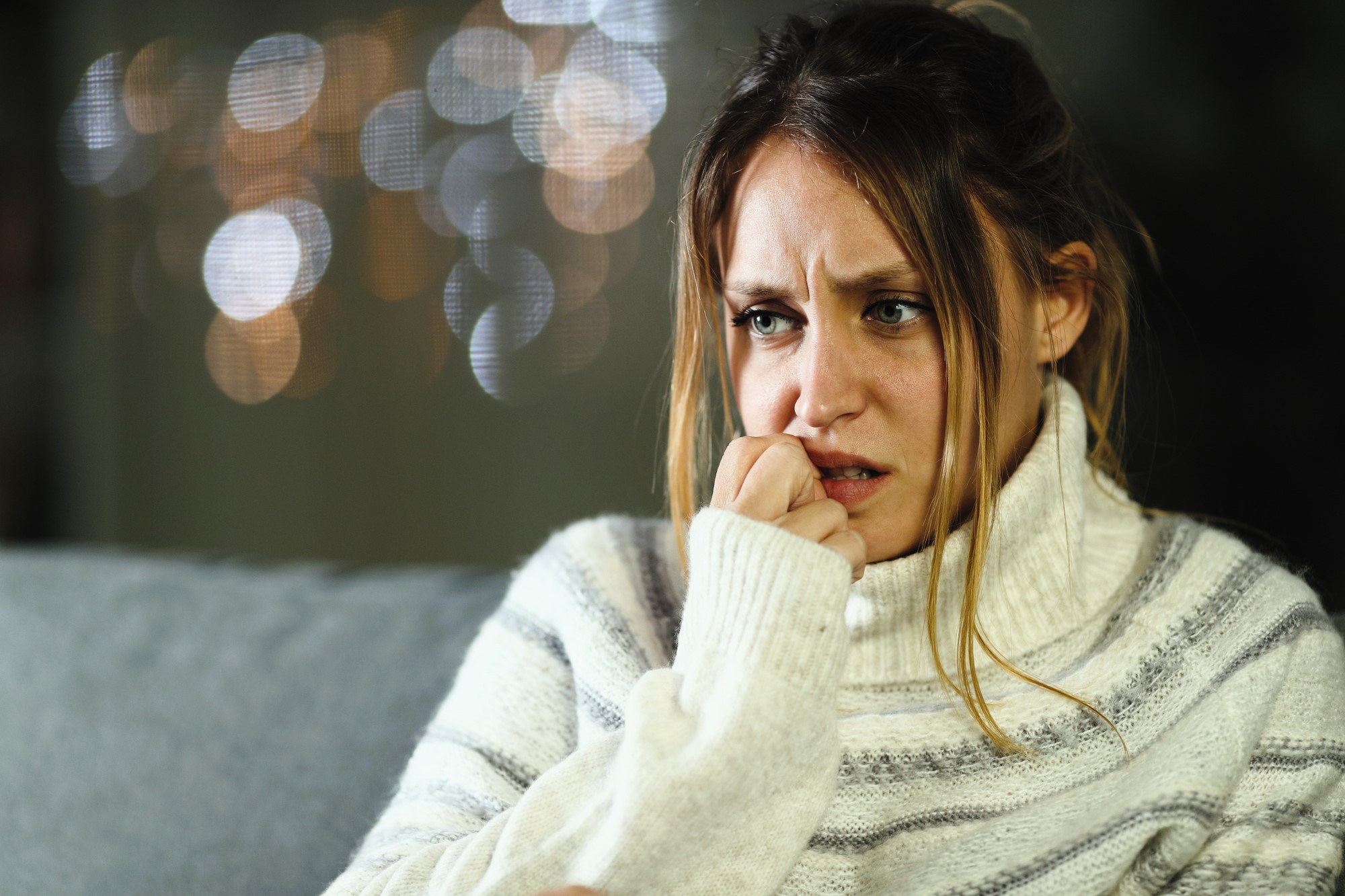 A woman wearing a striped turtleneck sweater appears worried or anxious, with her hand near her mouth. She is sitting indoors with out-of-focus lights in the background.