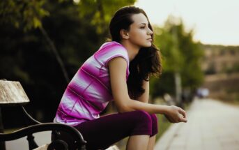 A woman sitting on a wooden bench outdoors, gazing thoughtfully into the distance. She is wearing a pink and white striped shirt and dark pants. Trees and a pathway are visible in the background.