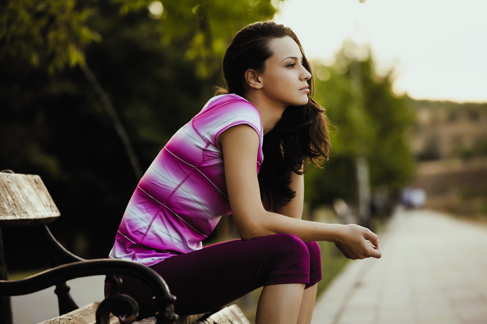 A woman sitting on a wooden bench outdoors, gazing thoughtfully into the distance. She is wearing a pink and white striped shirt and dark pants. Trees and a pathway are visible in the background.