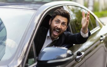 A man in a suit leans out of the window of a parked car, looking surprised or excited. He gestures with one hand, as if calling out or signaling to someone. The background includes greenery and an urban setting.