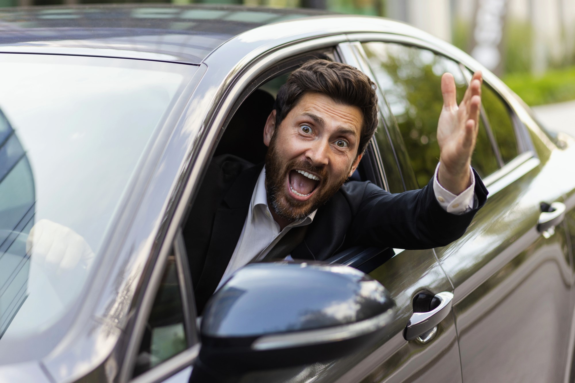 A man in a suit leans out of the window of a parked car, looking surprised or excited. He gestures with one hand, as if calling out or signaling to someone. The background includes greenery and an urban setting.