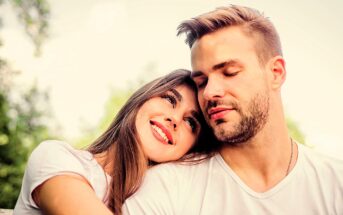 A woman with long brown hair leans her head on a man's shoulder while both smile gently. They are outdoors surrounded by blurred greenery and wear white shirts. The atmosphere is serene and affectionate.