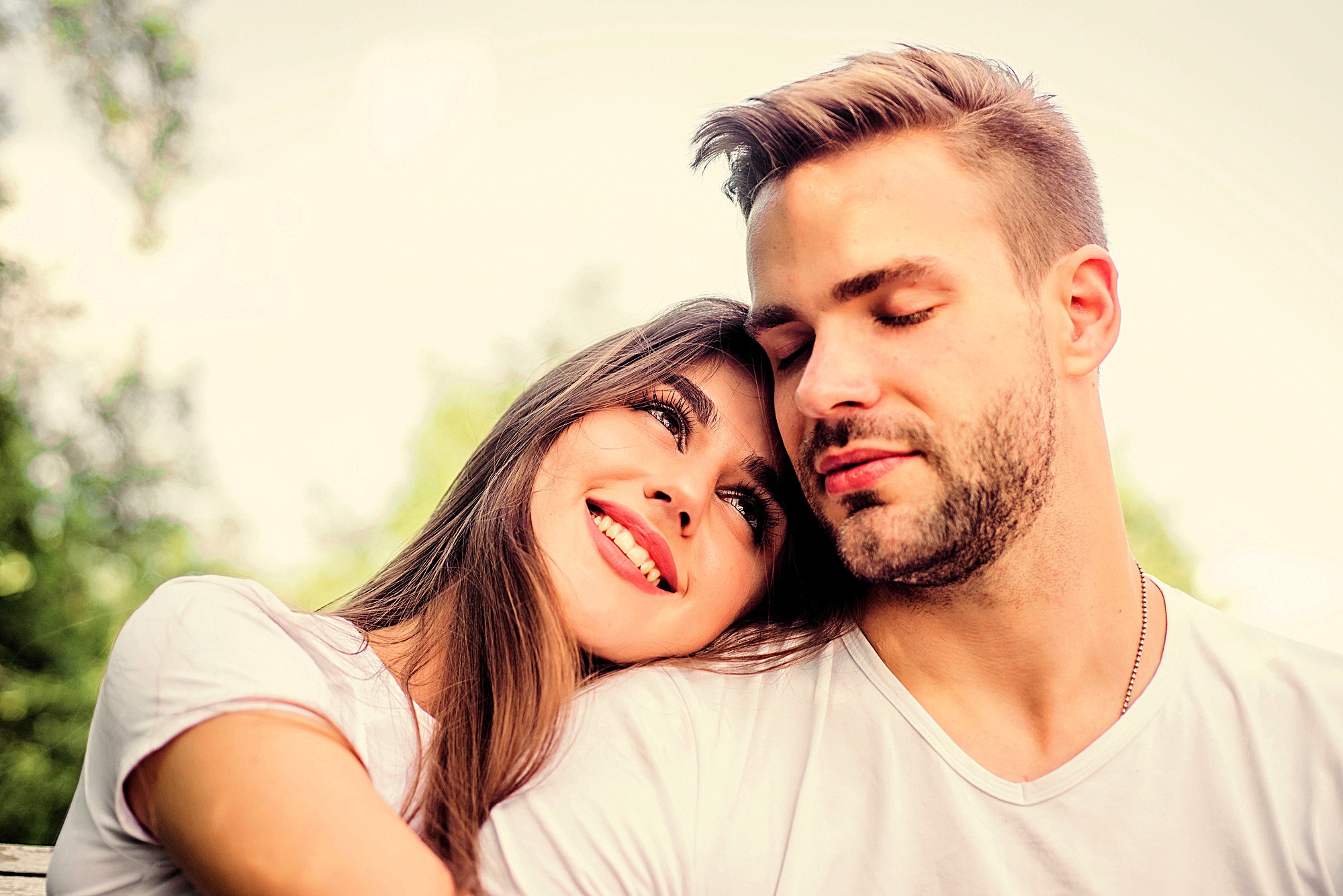 A woman with long brown hair leans her head on a man's shoulder while both smile gently. They are outdoors surrounded by blurred greenery and wear white shirts. The atmosphere is serene and affectionate.